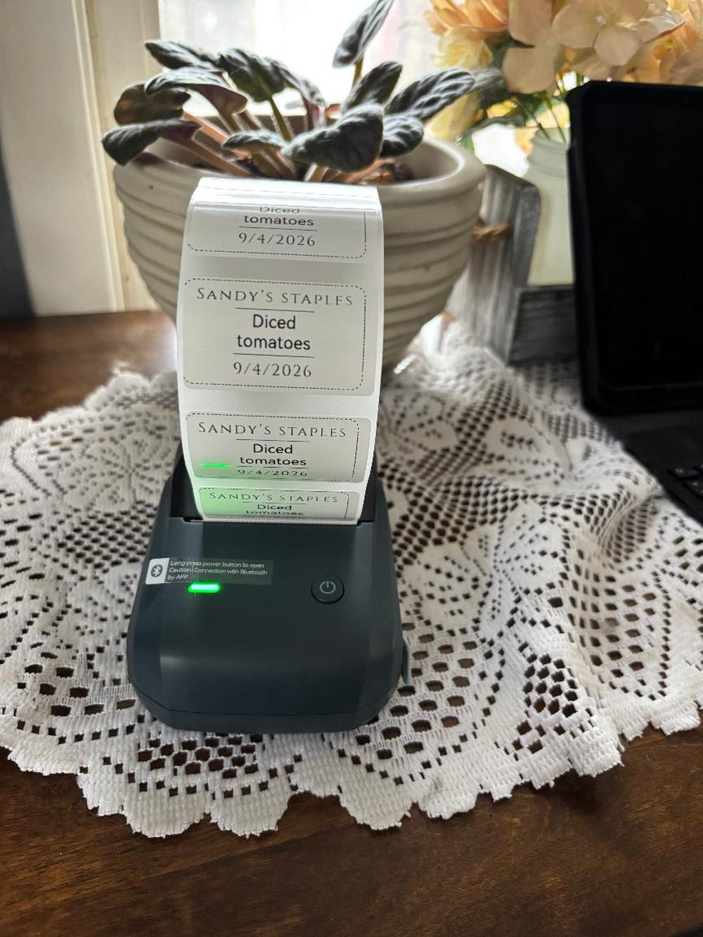 A small Bluetooth thermal label printer on a lace doily, printing a strip of labels reading SANDY’S STAPLES, Diced Tomatoes
