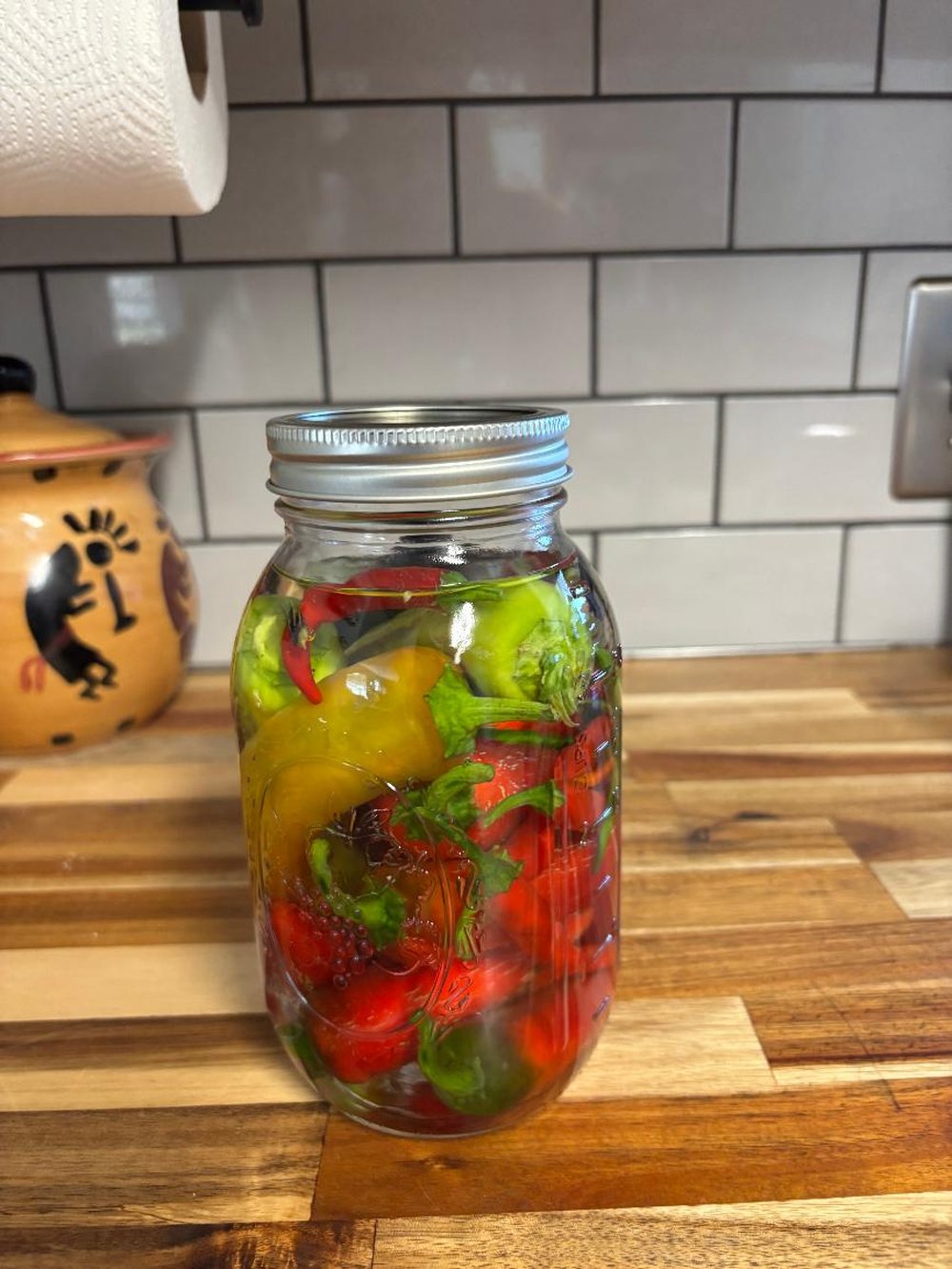 A mason jar of pickled peppers — red, green, and yellow — glowing like stained glass on a wooden counter